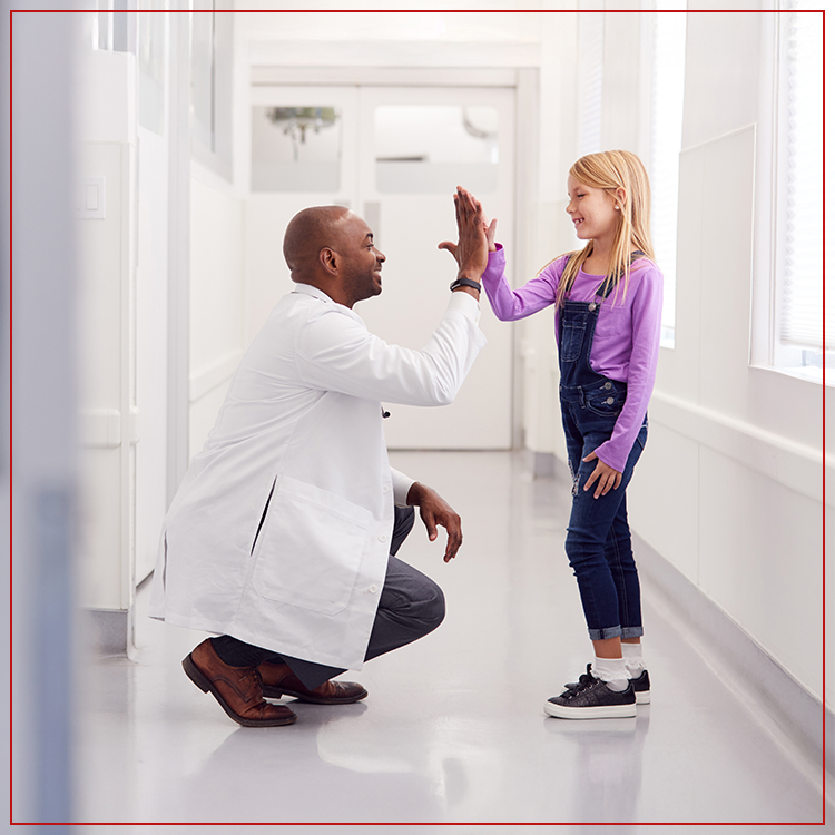 A doctor kneels in a hallway to give a high-five to a young girl in overalls and a purple shirt. Both are smiling, and the scene takes place in a bright, modern medical facility. | California Urgent Care Center