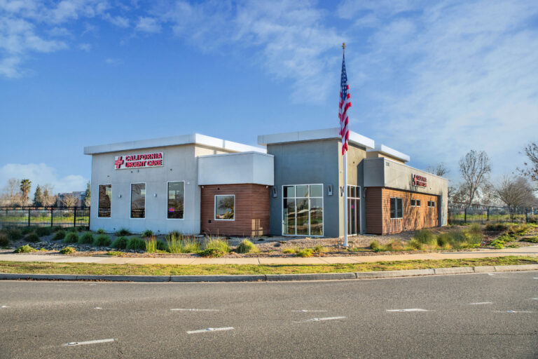 A modern medical building labeled California Urgent Care, with an American flag flying in front, surrounded by landscaping and a clear blue sky. The facility is located beside a road with marked lanes. | California Urgent Care Center