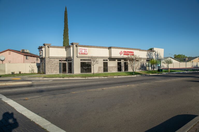 A street view of a California First Care building with a sign displaying open hours. The clinic is on a corner lot with a tree, shrubs, and clear blue sky in the background. | California Urgent Care Center
