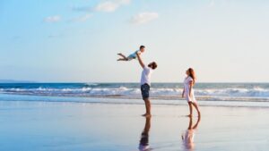 A man lifts a young child into the air while a woman smiles nearby. They are standing on a sandy beach with gentle waves and a blue sky in the background. | California Urgent Care Center