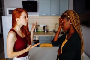 Two women in a kitchen are having an intense conversation; one gestures while talking, and the other covers her face with her hand, appearing upset or stressed. | California Urgent Care Center