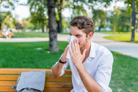 A man sitting on a park bench sneezes into a tissue, wearing a white shirt with a gray bag beside him, on a sunny day with green trees in the background. | California Urgent Care Center