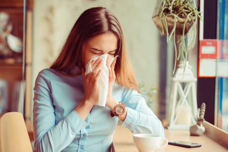 A woman sitting at a table in a café sneezes into a tissue. She has long brown hair and wears a light blue shirt and a watch. There is a cup, a smartphone, and a potted plant on the table. | California Urgent Care Center