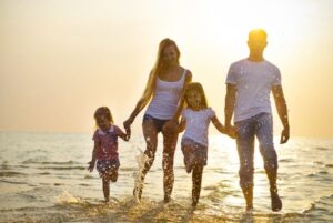 A family of four, two adults and two children, walks hand in hand along the beach at sunset, smiling and splashing in the shallow water. The sun shines brightly behind them. | California Urgent Care Center