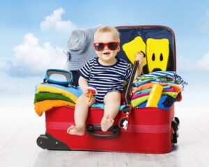 A smiling baby wearing red sunglasses and a striped outfit sits in an open red suitcase filled with colorful clothes, towels, and a sunhat, against a bright, blue sky background. | California Urgent Care Center