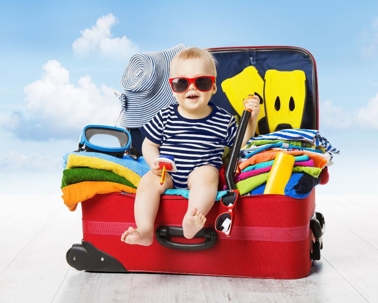 A smiling baby wearing red sunglasses and a striped outfit sits in an open red suitcase filled with colorful clothes, towels, and a sunhat, against a bright, blue sky background. | California Urgent Care Center