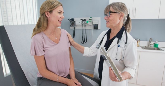A doctor with a clipboard talks to a smiling patient sitting on an exam table in a medical office, offering reassurance with a hand on the patients shoulder. Medical equipment is visible on the wall behind them. | California Urgent Care Center