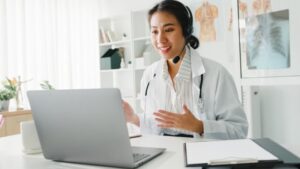 A female doctor wearing a headset and white coat sits at a desk, smiling and talking during a video call on her laptop. Medical charts and a clipboard are on the desk, with anatomical posters in the background. | California Urgent Care Center