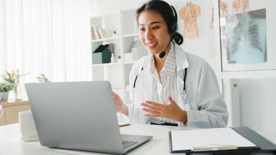 A female doctor wearing a headset and white coat sits at a desk, smiling and talking during a video call on her laptop. Medical charts and a clipboard are on the desk, with anatomical posters in the background. | California Urgent Care Center