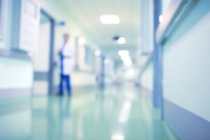 A blurred image of a hospital hallway with shiny floors, pale blue walls, and a person in a white coat standing in the distance. | California Urgent Care Center
