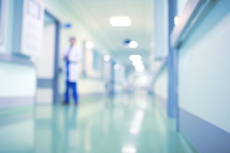A blurred image of a hospital hallway with shiny floors, pale blue walls, and a person in a white coat standing in the distance. | California Urgent Care Center