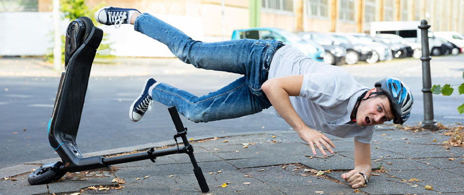 A young man wearing a helmet and jeans falls off an electric scooter onto a sidewalk, his arms extended to break his fall, with a surprised expression on his face. | California Urgent Care Center
