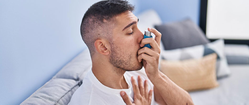 A man sitting on a couch uses an inhaler, holding it to his mouth with one hand while his other hand rests on his chest. He appears to be managing asthma or breathing difficulty. | California Urgent Care Center