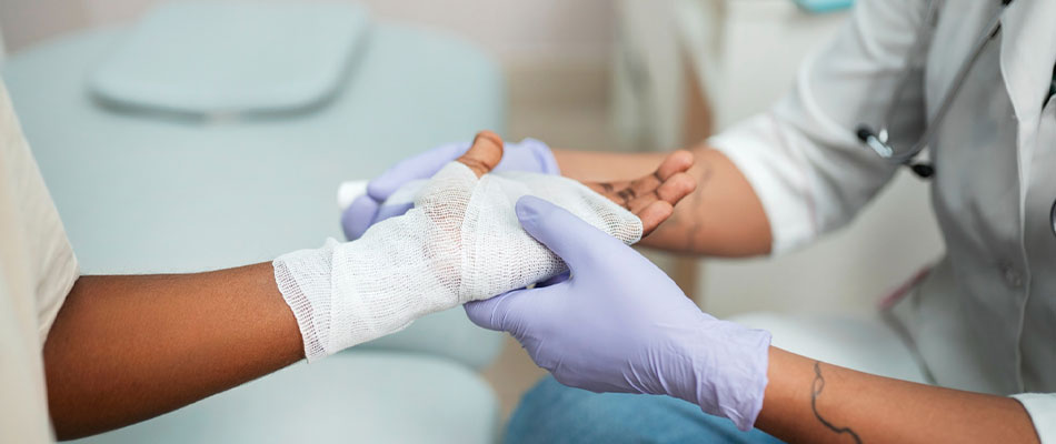 A healthcare professional wearing purple gloves wraps a bandage around a patients hand during a medical procedure in a clinical setting. | California Urgent Care Center
