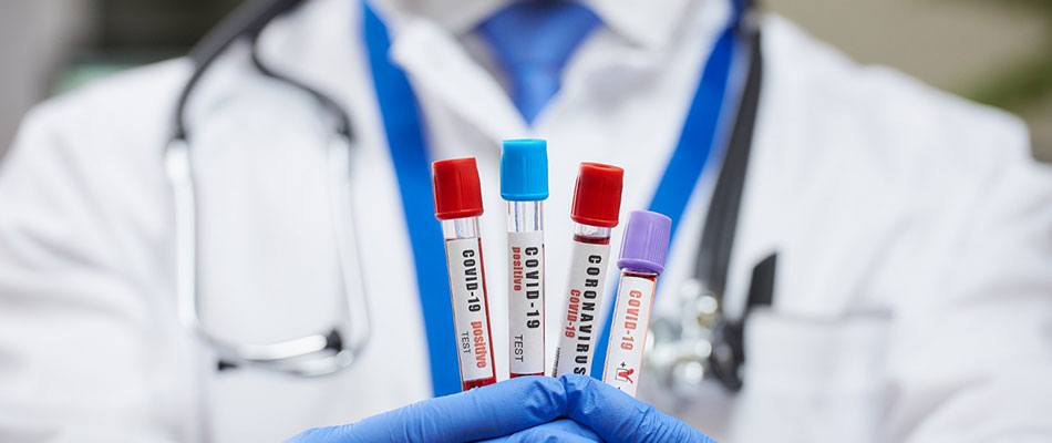 A doctor wearing a white coat, stethoscope, and blue gloves holds up four labeled COVID-19 test tubes, each with a different colored cap. | California Urgent Care Center