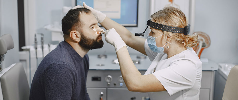 A female doctor wearing a face mask and gloves examines a seated mans nose with a medical instrument in a clinic setting. | California Urgent Care Center
