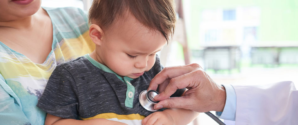 A doctor uses a stethoscope to listen to the chest of a young child sitting on an adults lap during a medical examination. | California Urgent Care Center