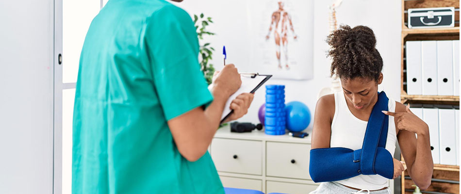 A woman with her arm in a blue sling sits in a medical office, looking at her injured arm while a healthcare professional in scrubs takes notes on a clipboard. | California Urgent Care Center