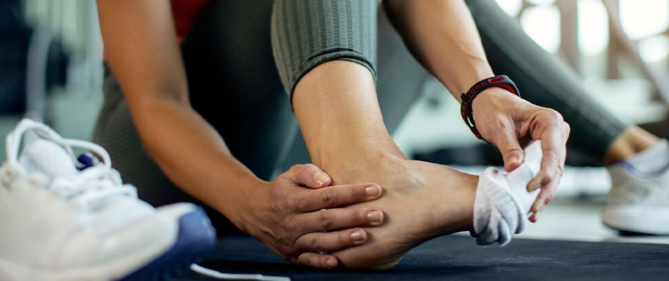 A person sitting on a mat holding their ankle and adjusting their sock, with one shoe off and workout equipment visible in the background, suggesting a possible foot or ankle injury during exercise. | California Urgent Care Center