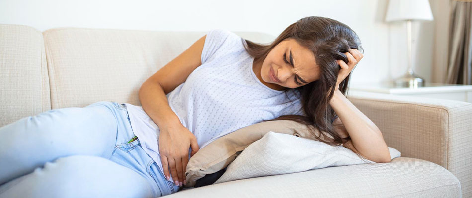 A young woman lies on a couch, clutching her abdomen with a pained expression, possibly experiencing stomach pain or cramps. | California Urgent Care Center